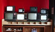 Premium stock video - Stacks of vintage televisions with few turned-on on wooden rack against red wall