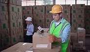 Free stock video - Warehouse worker wearing a helmet and reflective vest seals a box with duct tape, in the blurred background a woman is placing cardboard boxes on the shelf