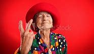 Portrait of funny, elderly mature woman, 80s, giving peace sign gesture with no teeth and red hat, isolated on purple background studio. Concept of youthful old lady with copy space