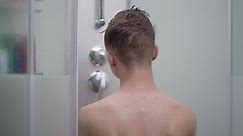 Young boy bathing under a shower at home - back view. Beautiful teen boy taking shower and washing in the bathroom. Happy child washing head, face and body with water.