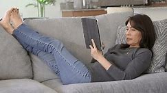 Attractive mature woman in denim jeans bare feet up wiggling toes relaxing shopping online using iPad mobile tablet excited happy at successful internet purchase waving e-commerce credit card.