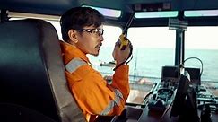 Filipino deck Officer on bridge of vessel or ship wearing coverall during navigaton watch at sea . He is speaking on GMDSS VHF radio, communication between vessels.