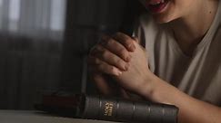 Woman prays with her hands crossed over Bible for God blessing in dark room. Faith and love in God.Christian prayer over Bible. Woman worshiping Christian God. Catholic faith.Girl prays in an epidemic