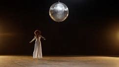 Charming African American Woman with Fluffy Afro Style Hairs in White Jumpsuit Wearing Roller Skates Rides Around on the Stage Under Shiny Disco Ball. Cinematic Slow Motion Shot