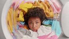 Sad Afro American woman poses in washing machine drum with clothes for washing falling on her has much housework feels tired loads washer with laundry. Frustrated housekeeper does laundering