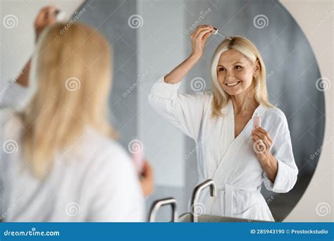 Mature Woman Applying Hair Repair Serum While Standing Near Mirror In