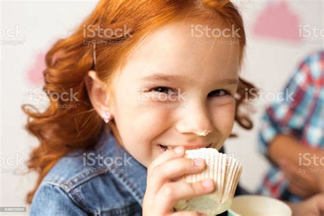 Cute Little Redhead Girl Eating Sweet Cupcake And Looking At Camera