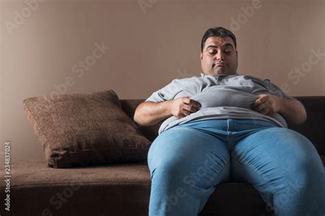Obese Man Sitting On Sofa Holding His Belly Fat With Both Hands And