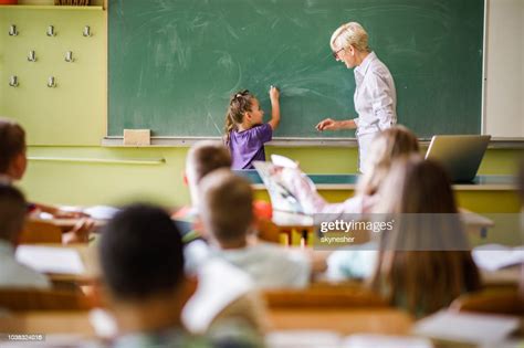Senior Female Teacher Teaching Schoolgirl In Front Of Blackboard At