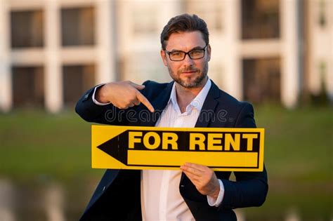 Successful Real Estate Agent In A Suit Holding For Rent Sign Near New