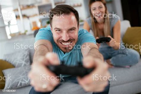 Husband And Wife Playing Video Game With Joysticks In Living Room Stock