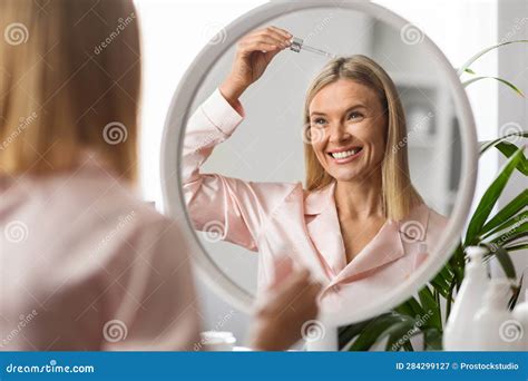 Mature Woman Applying Repair Hair Serum While Standing Near Mirror At