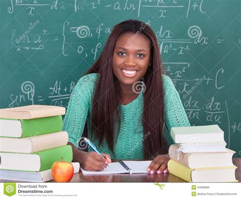 Confident Female Teacher Writing In Book At Classroom Desk Royalty Free