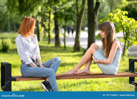 Two Young Girls Friends Sitting On A Bench In Summer Park Chatting