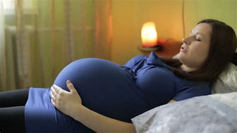 a pregnant woman in a dress is sitting on a bed and then lying down and stroking big belly a