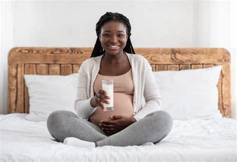 happy pregnant african woman drinking milk while sitting on bed at home
