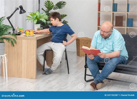 Dad Is Sitting On The Sofa Reading A Book To His Son And He Is Sitting