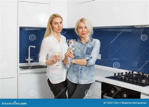 Two Blonde Girlfriends Drink Wine In The Kitchen And Laugh Two Adult
