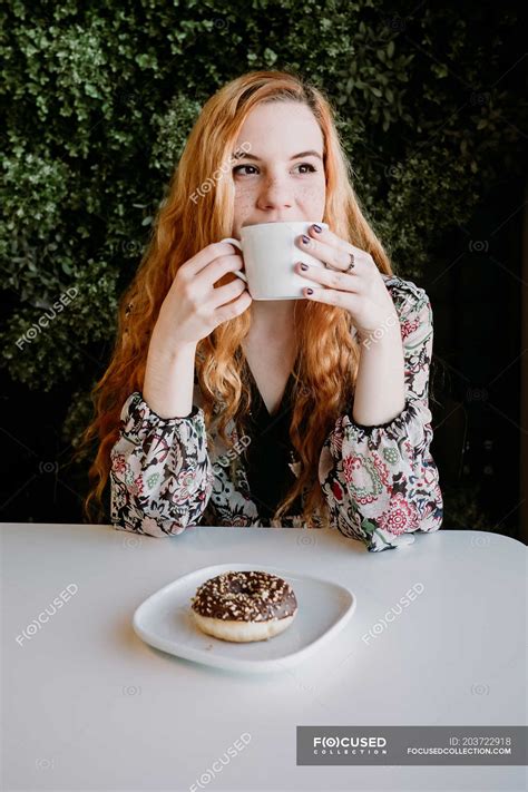 smiling redhead woman with cup sitting and doughnut sitting against