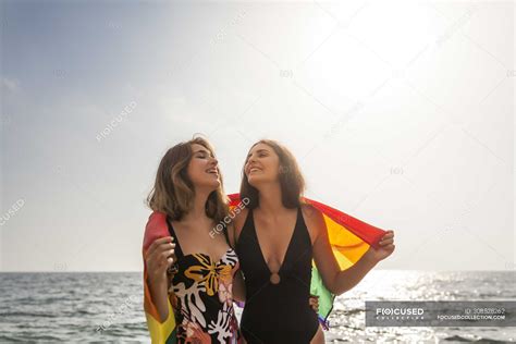 Lesbian Couple Standing On Beach Hugging Wrapped In Colorful Flag Of
