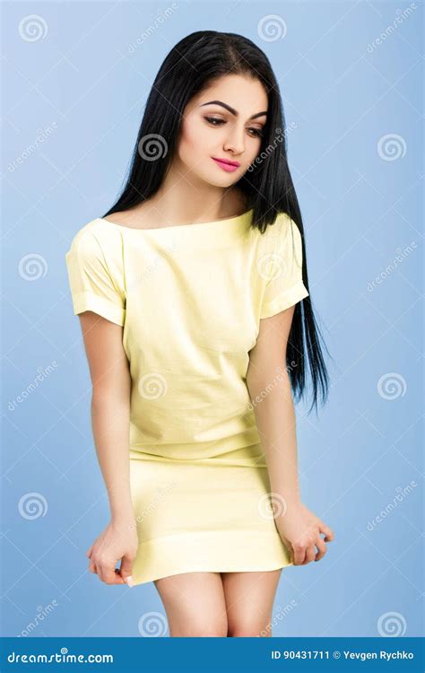 Shy Young Brunette Woman Adjusts Her Yellow Dress On Blue Background