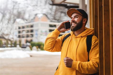 Bearded Hipster Man Tourist In Yellow Hoodie And Cap Stands Outdoors