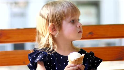 little blonde girl eats licks an ice cream cone on a park bench