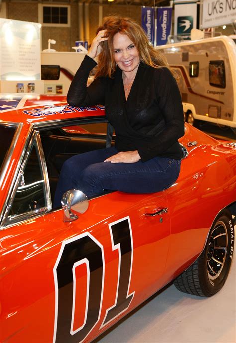 a woman sitting on the hood of an orange car in a showroom with other cars
