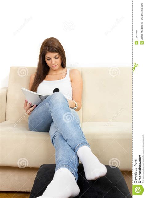 muchacha en la camiseta blanca y vaqueros que leen la revista en el sofá fotografía de archivo