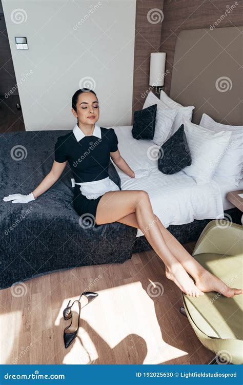 Maid In White Apron And Gloves Sitting On Bed With Closed Eyes In Hotel