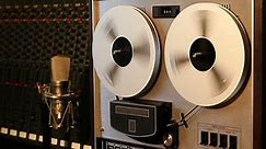 Stereo reel to reel tape recorder rewinding and fast forwarding with studio mixer and microphone in the background.