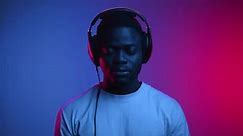 Portrait of African American Man in Headphones Listening Music in Colorful Neon Light. Calm Hobby of 20s Black Person Hearing Broadcast of Hip Hop Record. One Technology Lover Uses Portable Head Set