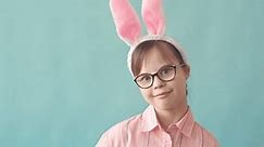 Chest up studio portrait of 12 year old Caucasian girl with down syndrome wearing bunny ears smiling at camera standing on light blue background