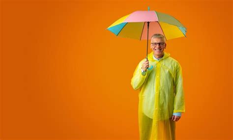 Senior man wearing yellow plastic overcoat and holding rainbow umbrella ...
