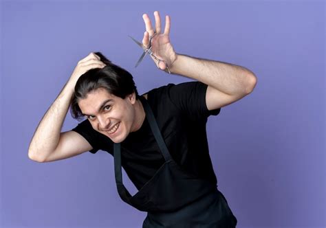 Smiling young handsome male barber in uniform holding scissors and ...
