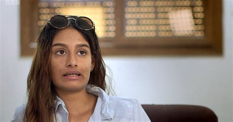 Shamima Begum leaning against a door frame. She has long dyed brown hair and is wearing sunglasses with a light grey top underneath a sweatshirt jacket