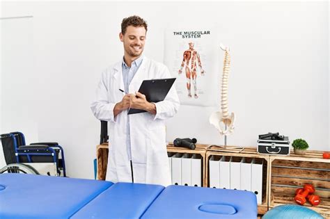 Young hispanic man wearing physiotherapist uniform writing on clipboard ...
