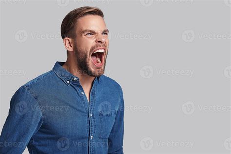 Furious man. Angry young man screaming while standing against grey ...
