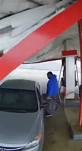 CCTV footage captures the terrifying moment a gas station canopy suddenly collapses without warning. Filmed from a security camera above the pumps, the clip shows a silver sedan parked below as a man stands beside it, completely unaware of the danger overhead. In an instant, the entire metal roof buckles and crashes down with massive force, crushing part of the car and the fuel pump. Large panels slam into the ground as dust and debris explode across the concrete. The man reacts just in time, cr