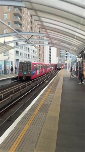 The video shows a train platform at a train station. A red and white train is on the tracks, approaching the platform. There are buildings in the background. Several people are visible on the platform, waiting for the train. The platform is covered by a translucent roof. The train appears to be a light rail or metro train. in London, United Kingdom | Collective Memory