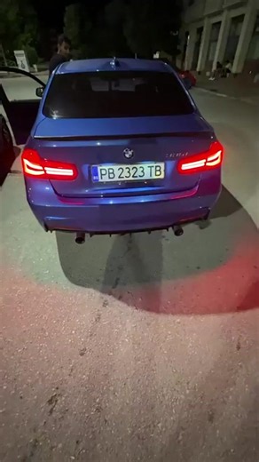 The video shows the rear of a blue BMW sedan parked on a street at night. The car's brake lights are illuminated, casting a red glow on the asphalt. The license plate reads "PB 2323 TB" and displays the Bulgarian flag. Two exhaust pipes are visible at the rear of the car. In the background, a person can be seen standing near the open driver's side door of the car. Further in the background, there are buildings, trees, and other cars, suggesting an urban or suburban setting. The lighting indicate