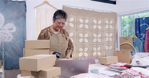 Senior woman entrepreneur stands near laptop, scans parcel barcode, moves carton into stack, checks order management list, prepares e-commerce shipment queue inside home studio workshop home studio.