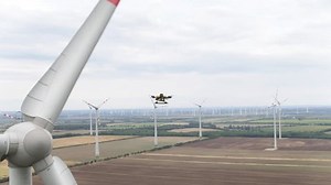 A drone flies amongst rotating wind turbines in an electricity generating farm, as it surveys and inspects the condition of the structures