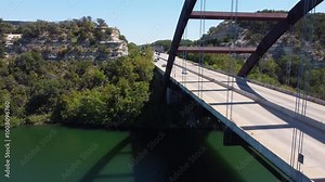 An aerial shot captures the iconic Pennybacker Bridge in Austin, Texas, with cars driving across it, showcasing the beautiful landscape and water below.