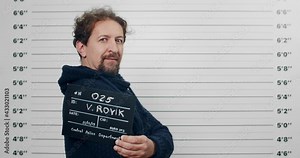 Side profile mugshot of male arrested in 40s turning head and looking to camera. Criminal guy with blue eyes holding sign for photo while standing in front of police metric lineup wall.