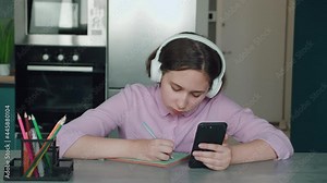 Cute child girl wearing headphones studying online in mobile app by video call with remote teacher, tutor. School kid holding phone doing homework using application, watching class learning at home.