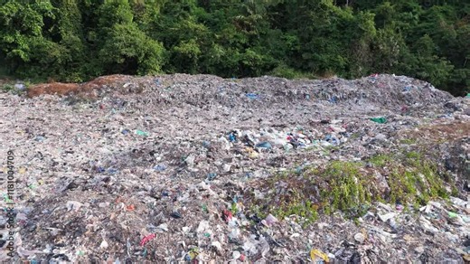 Aerial drone footage captures a large garbage landfill site on a tropical island in Thailand, highlighting the environmental challenges faced in waste management. Surrounded by lush greenery