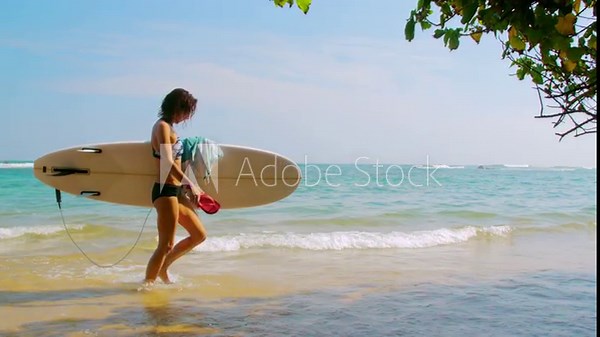 Relaxed woman with surfboard holds slates walking in warm ocean water. Female surfer enjoys light breeze after catching waves