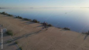 Young female athlete riding a road bicycle on gravel road with a beautiful calm blue lake and behind. Back side view tracking drone footage in slow motion.