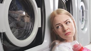 Close-up of a young woman with a sad expression sitting by the washing machine in a public laundry.